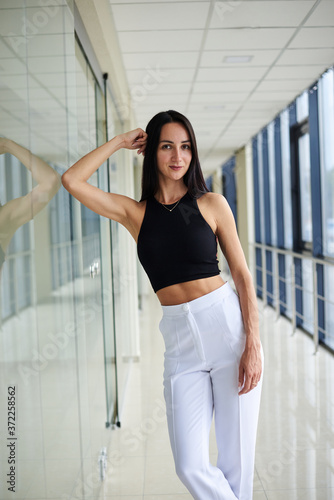 Young brunette woman, wearing white pants and black top, standing in light passageway with huge windows, posing, leaning on glass wall. Businesswoman on a break. Female portrait photography. Free Download Young brunette woman, wearing white pants and black top, standing in light passageway with huge windows, posing, leaning on glass wall. Businesswoman on a break. Female portrait photography. - GFXLoad