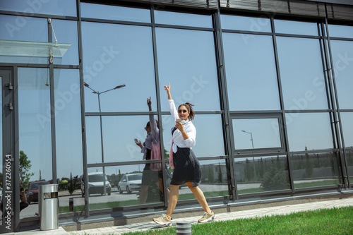 Young woman walking against glass' wall in airport, traveler with small baggage, influencer's or blogger's lifestyle. Caucasian female model with gadgets before starting her vacation, registration.