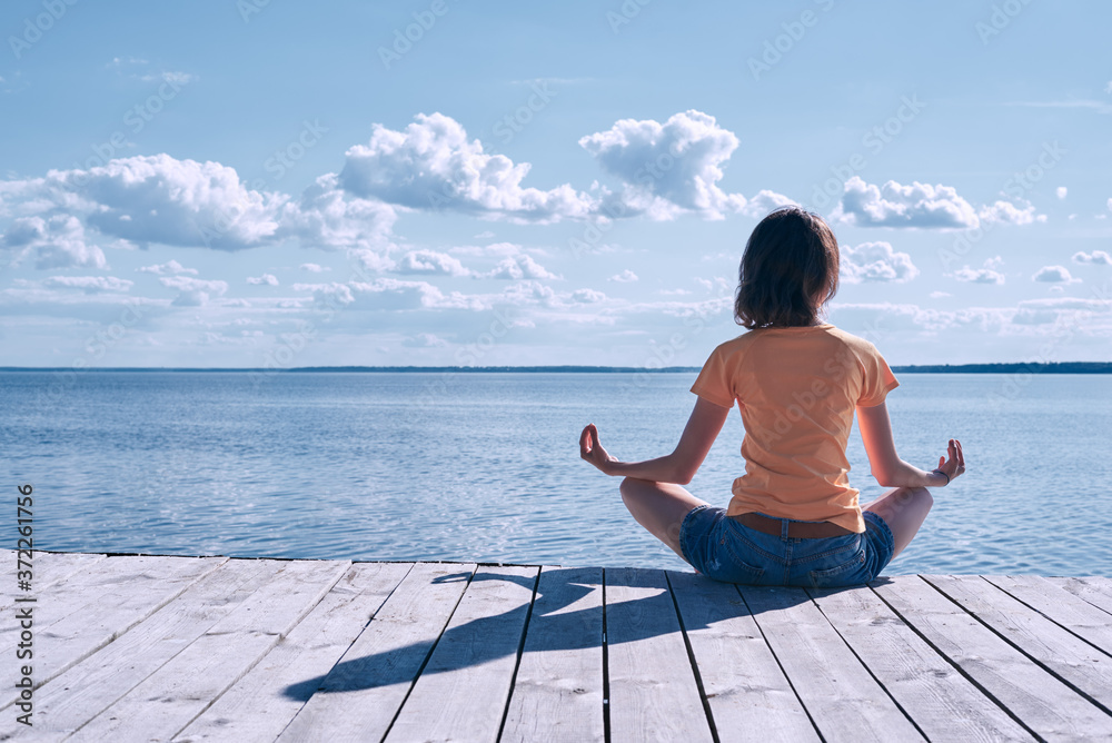 Side view of A young pretty woman enjoys the rest, sitting on the shore of the river on a wooden seafront in lotus position