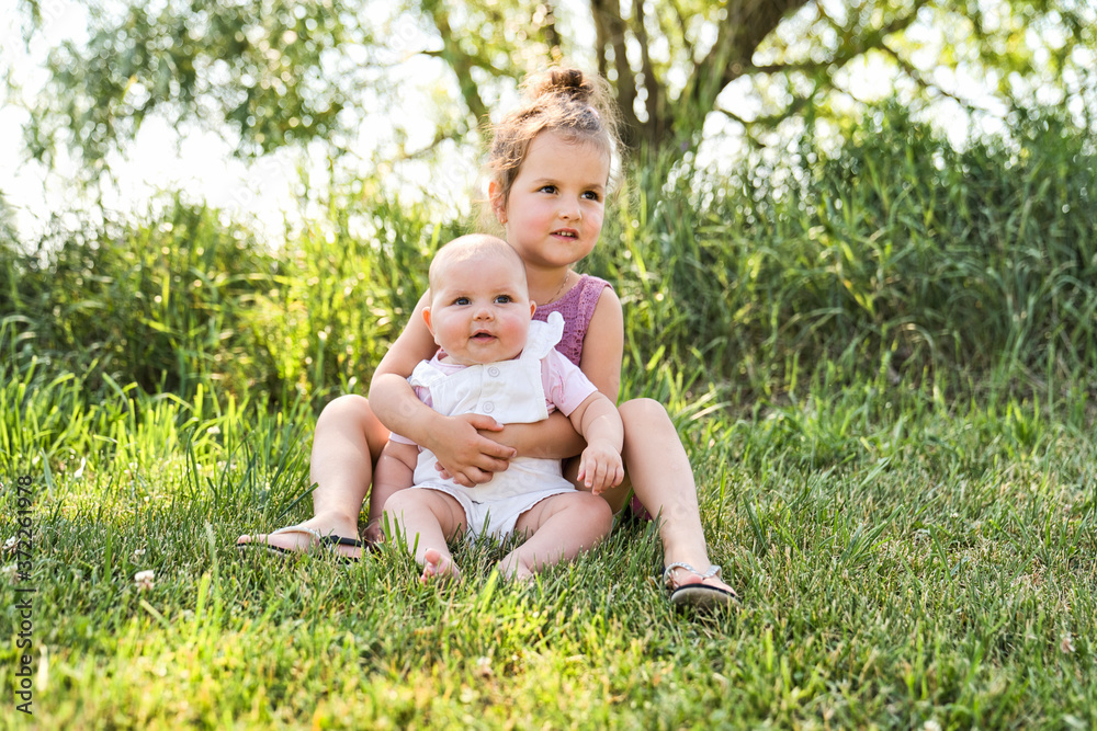 Happy adorable chubby baby girl sitting on the grass with her daughter sister Happy adorable chubby baby girl sitting on the grass with her daughter sister