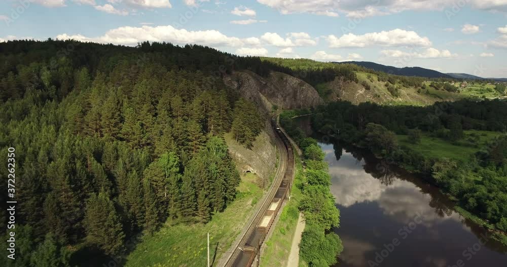 Freight long train carries with empty carriages an electric locomotive by two-sided Trans Siberian railways under the rock and near mountains river / Aerial drone wide view at summer sunny day Freight long train carries with empty carriages an electric locomotive by two-sided Trans Siberian railways under the rock and near mountains river / Aerial drone wide view at summer sunny day