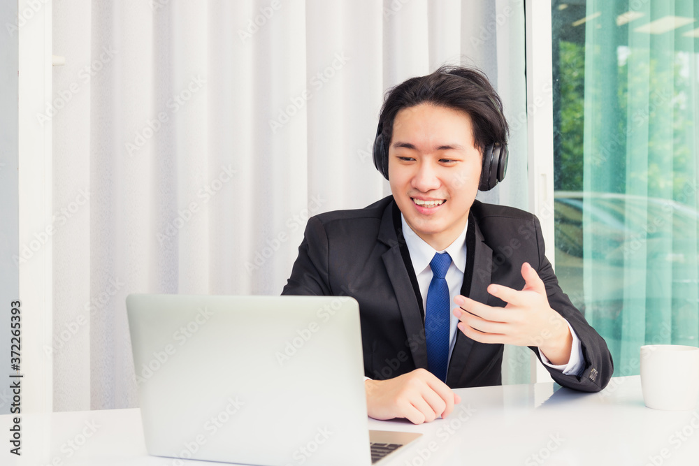 Work from home, Asian young businessman smile wearing headphones and suit video conference call or facetime on desk and raise his hand to explain to colleagues at home office Work from home, Asian young businessman smile wearing headphones and suit video conference call or facetime on desk and raise his hand to explain to colleagues at home office