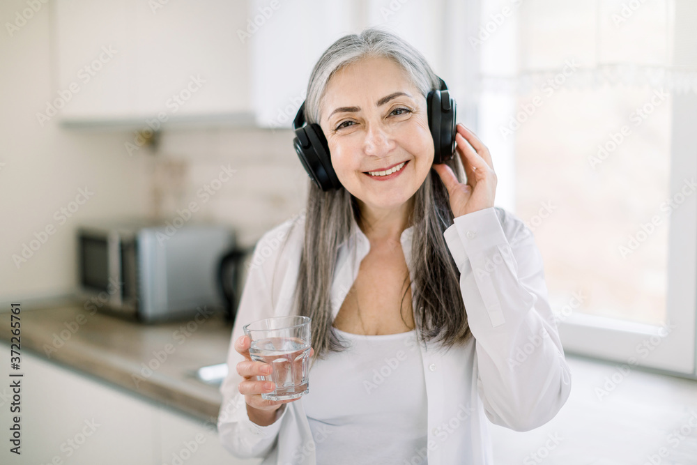 Portrait of happy smiling mature granny with long gray hair, standing in modern light kitchen with glass of water, listening to the music or audio book using headphones Portrait of happy smiling mature granny with long gray hair, standing in modern light kitchen with glass of water, listening to the music or audio book using headphones