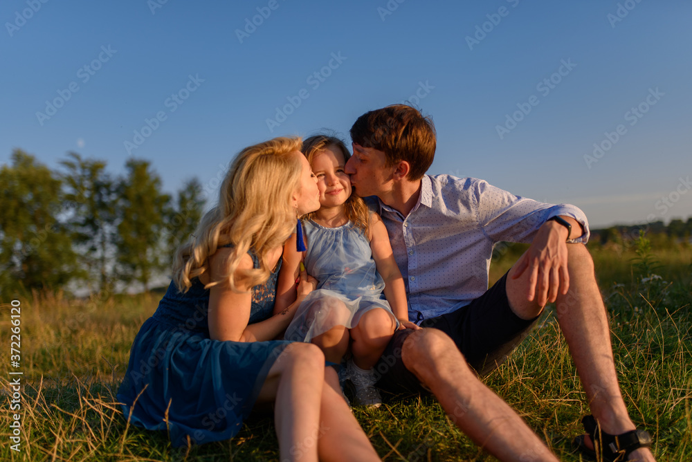 Dad mom and daughter are sitting together on the grass. Parents kiss their daughter on the cheeks. Dad mom and daughter are sitting together on the grass. Parents kiss their daughter on the cheeks.