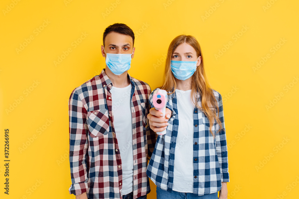man and a woman in a medical protective mask on their face, a woman checks the temperature with a digital thermometer, standing on a yellow background, coronavirus