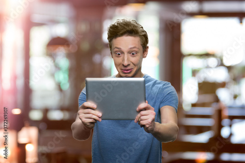 Stunned teen boy looking at digital tablet. Shocked teen guy using pc tablet against blurred background.