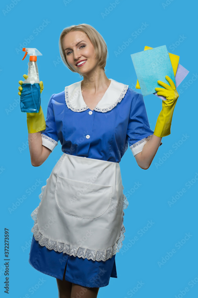 Cheerful maid in uniform holding rags and detergent. Smiling house maid posing with cleaning equipment on color background. Cheerful maid in uniform holding rags and detergent. Smiling house maid posing with cleaning equipment on color background.