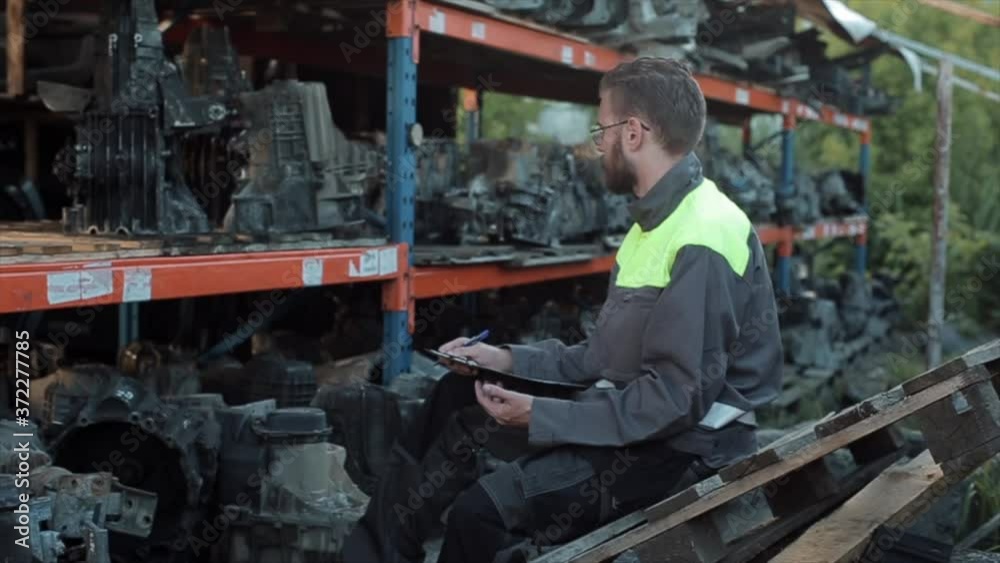 A responsible young bearded mechanic with glasses sits in front of racks of spare parts in a workshop on a pallet and taking notes in a notebook. Side view A responsible young bearded mechanic with glasses sits in front of racks of spare parts in a workshop on a pallet and taking notes in a notebook. Side view