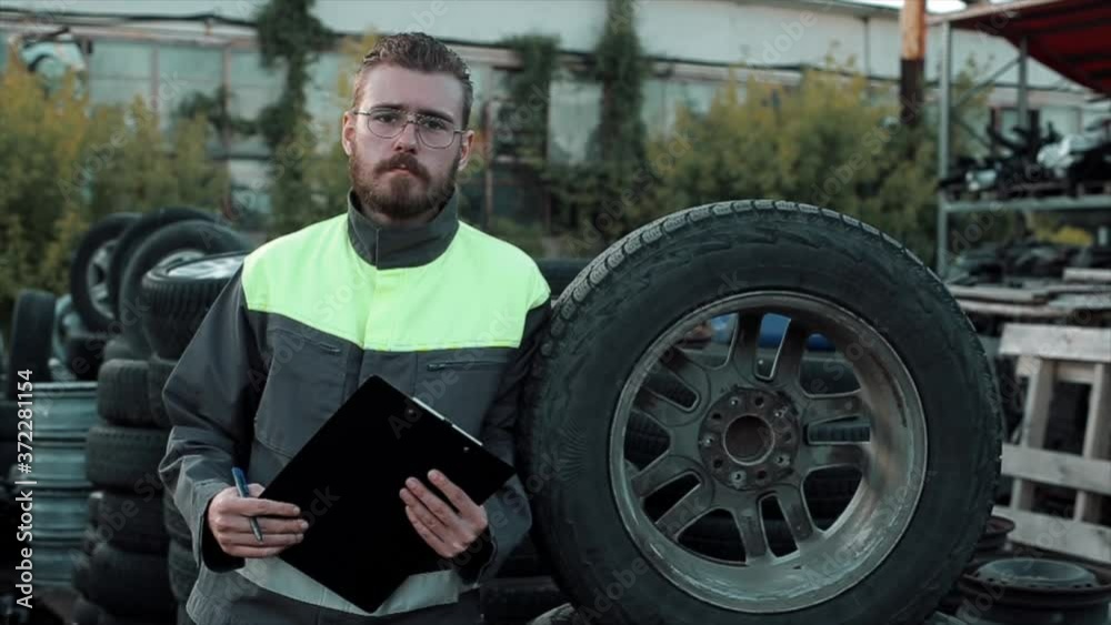 Portrait of a young bearded mechanic in glasses in gray overalls who stands next to a wheel in a car service warehouse with a notebook in his hands Portrait of a young bearded mechanic in glasses in gray overalls who stands next to a wheel in a car service warehouse with a notebook in his hands
