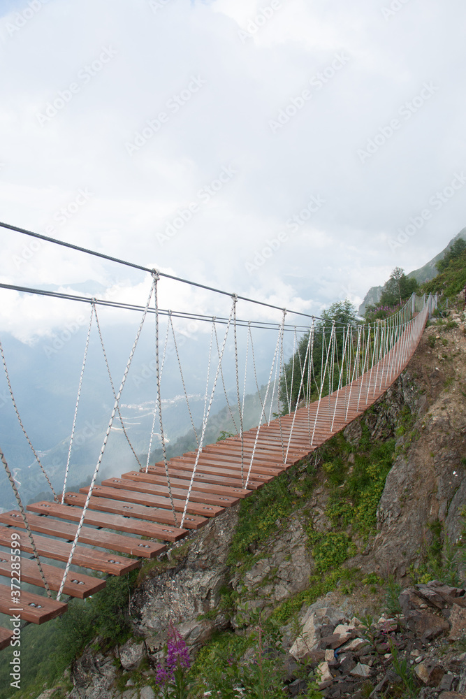 Obraz premium Skypark. An extreme, wooden rope suspension bridge over the chasm between two mountain peaks. Around the cloud. Sunny summer day. Vertical photo.