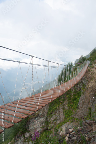 Skypark. An extreme, wooden rope suspension bridge over the chasm between two mountain peaks. Around the cloud. Sunny summer day. Vertical photo.