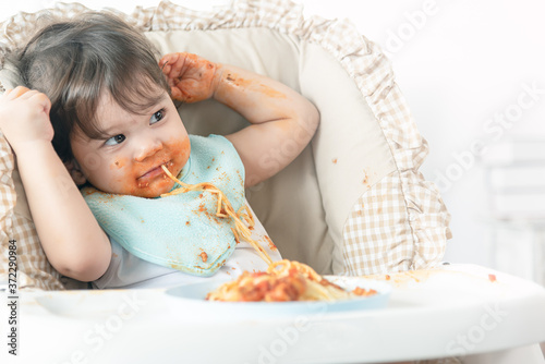Messy one year baby girl eating pasta.  Parents leave lovely infant girl, enjoying eating meal alone. Curious baby learns eating herself.