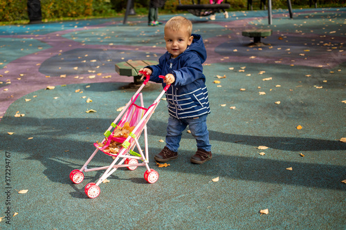 cute toddler kid carries yellow leaves in baby stroller in the autumn park and smiles at the camera