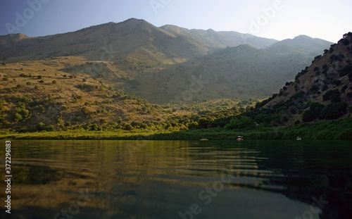 Idyllic peaceful view of mountain lake. Calm water surrounded by green mountains with bushes and trees, bright blue sky