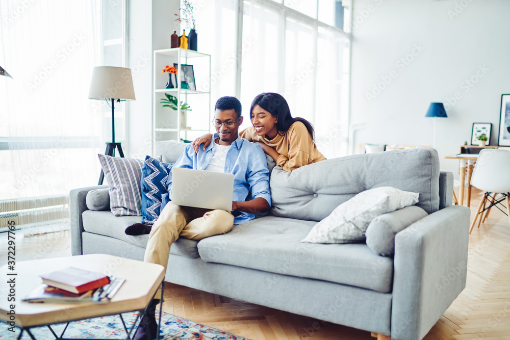 Positive african american woman watching video on laptop computer with boyfriend spending free time at home, dark skinned couple in love making online shopping together on netbook in living room.