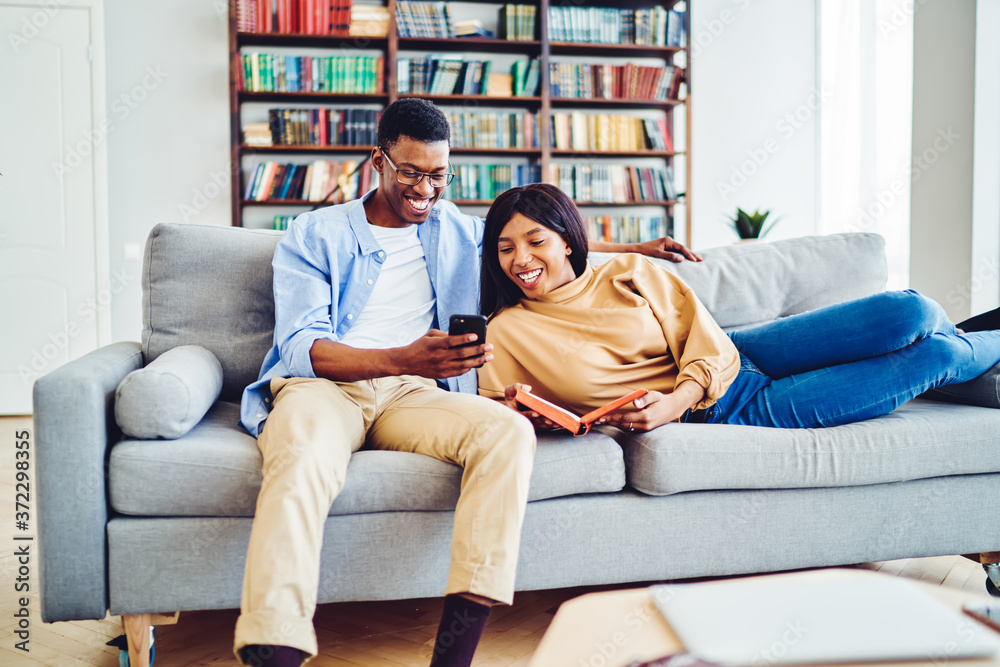 Cheerful dark skinned man showing funny video on smartphone to his girlfriend resting together at home interior, happy young marriage laughing at photos from social networks using mobile phone rest.