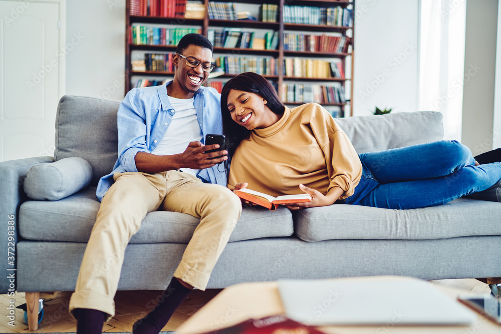 Smiling dark skinned couple in love having fun together resting at comfortable sofa in bright living room,happy african american guy showing video to girlfriend on mobile phone laughing at home. Smiling dark skinned couple in love having fun together resting at comfortable sofa in bright living room,happy african american guy showing video to girlfriend on mobile phone laughing at home.