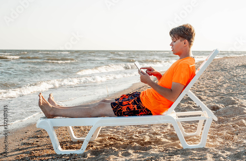 A guy in a T-shirt and shorts holds a tablet while lying on a sun lounger by the sea. photo