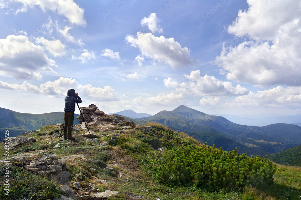 Young photographer man standing on top of cliff taking pictures of the ridge mountain range of Chernogor in Ukraine. Carpathian summer mountains