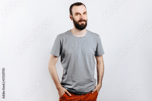 A handsome brutal brunette man with a beard in a gray shirt smiles and looks at the camera, holds his hand in his pockets. Place for advertising on a gray background.