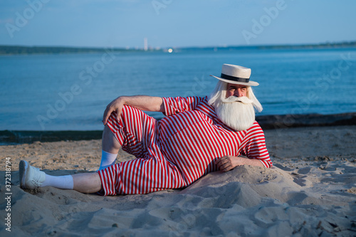 An elderly man in a striped retro swimsuit sunbathes on the beach. An old gray-haired bearded man in a hat lies on the sand by the sea.