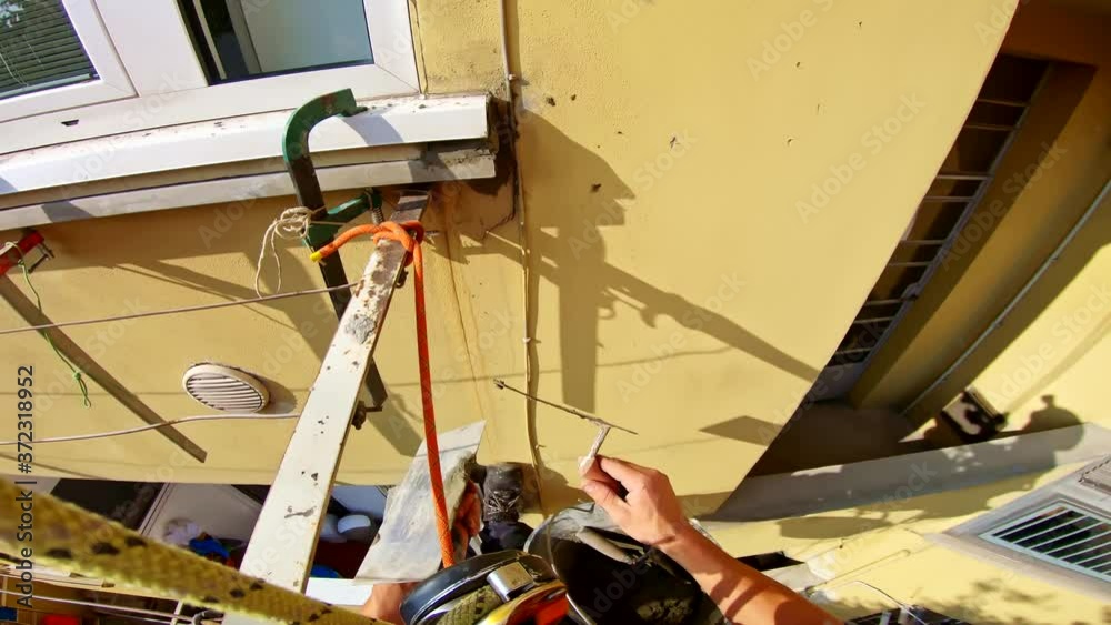 Construction home worker applying cement on wall in rope access gear, point of view