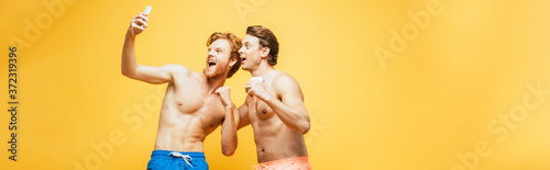 horizontal image of two shirtless friends grimacing and showing clenched fists while taking selfie on smartphone isolated on yellow