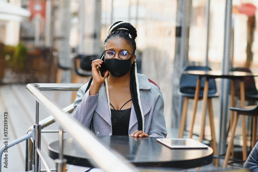 Fototapeta premium young pretty african american women in a medical protective mask outside in cafe. african girl with tablet and phone.