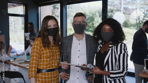 COVID-19 safety at workplace. Happy young multiethnic colleagues look at camera wearing face masks at modern office.