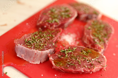 Five raw meat pieces of steak on a red cutting board, sprinkled with rosemary and coarse salt.