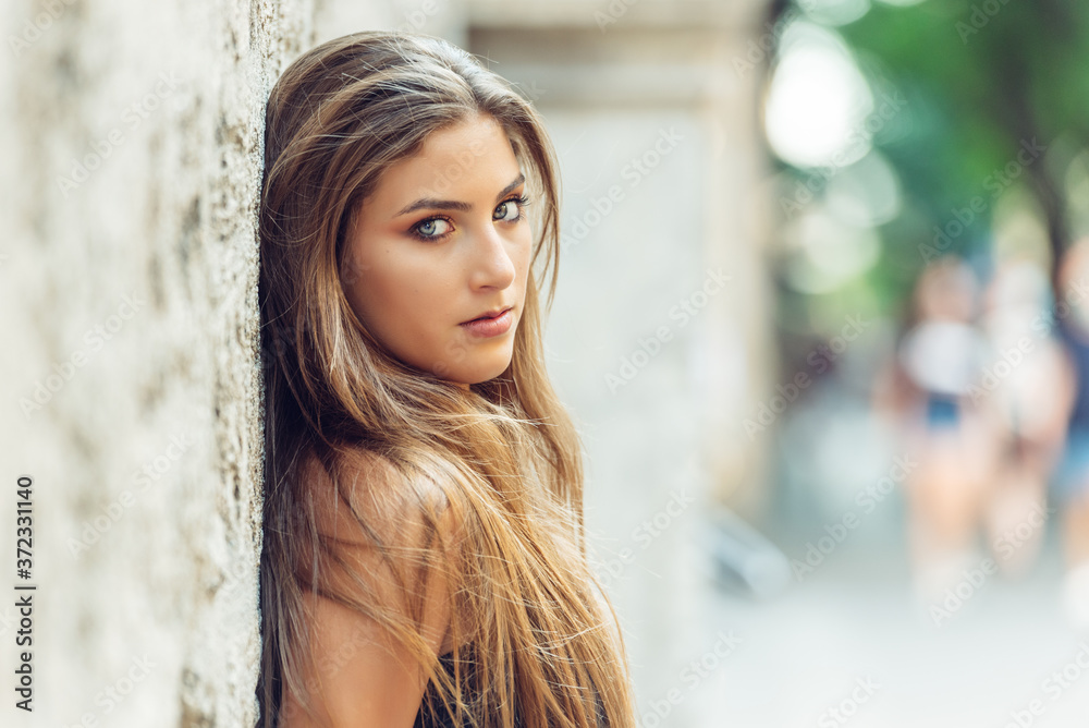 side view portrait of blond young woman with light eyes looking at the camera, with her back leaning against stone wall. Selective focus side view portrait of blond young woman with light eyes looking at the camera, with her back leaning against stone wall. Selective focus