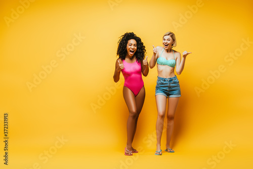 full length view of excited interracial women in summer outfit showing thumbs up while looking at camera on yellow
