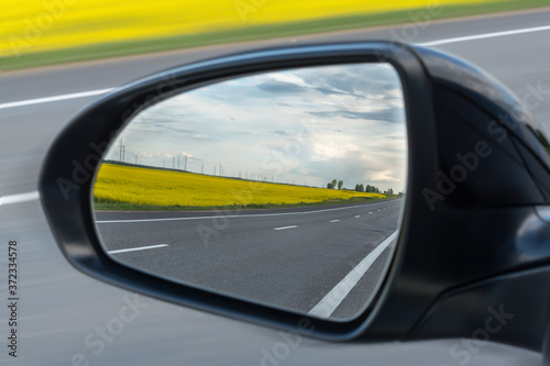Asphalt road, yellow flowering fields and cloudy sky are reflected in the car mirror