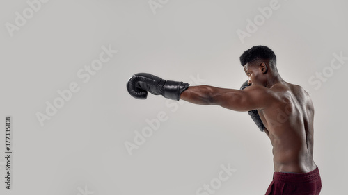 Portrait of young muscular african american male boxer looking aside, wearing boxing gloves, punching, standing isolated over grey background. Sports, workout, bodybuilding concept