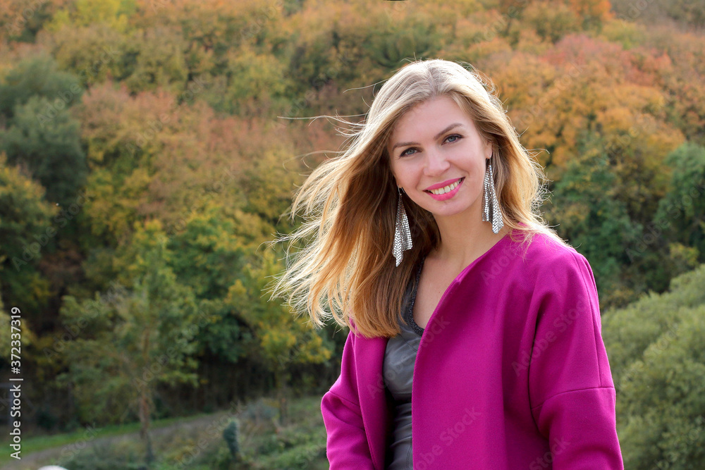 Happy smiling  blonde woman, wear coat in autumn time, young woman in fuchsia color design coat, trees of fall colors, 