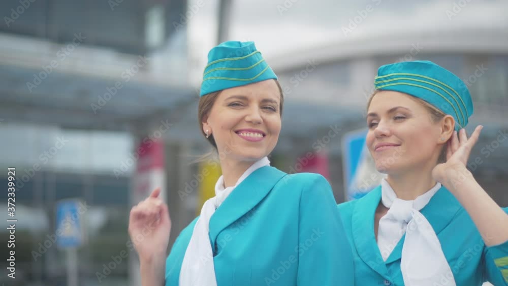 Two gorgeous women in stewardess uniform posing at the background of airport building. Portrait of smiling confident Caucasian flight attendants standing outdoors looking at camera and smiling. Two gorgeous women in stewardess uniform posing at the background of airport building. Portrait of smiling confident Caucasian flight attendants standing outdoors looking at camera and smiling.
