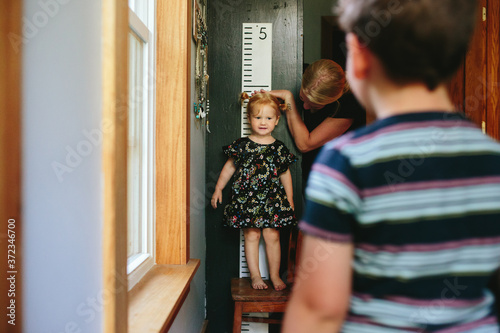 little girl stands on chair to be taller while mom measures her height