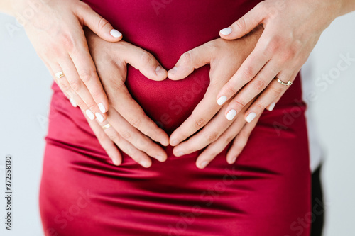Concept of pregnancy and motherhood, unity of the family. Husband and wife together. A man holds the belly of his beloved pregnant woman. She puts her hands on top of his hands. Heart shaped fingers