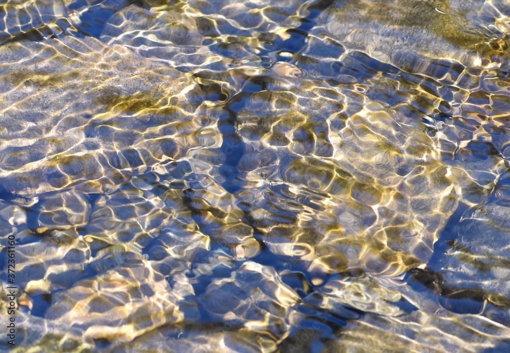 Peaceful and rippled surface of a stream, with waves and patterns of sunlight, brown rocky bottom visible underneath.  