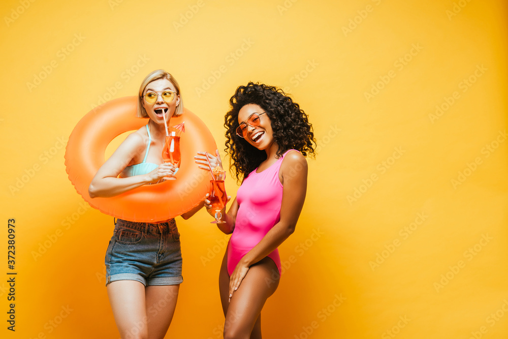 excited interracial women with swim ring and cocktail glasses posing on yellow excited interracial women with swim ring and cocktail glasses posing on yellow