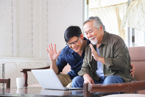 Elder Asian man and his son using video call from computer to say hi to the rest of his family in his cozy retirement home with copy space