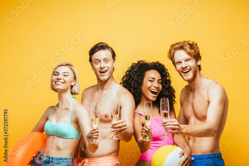 excited multicultural friends looking at camera while holding champagne glasses isolated on yellow