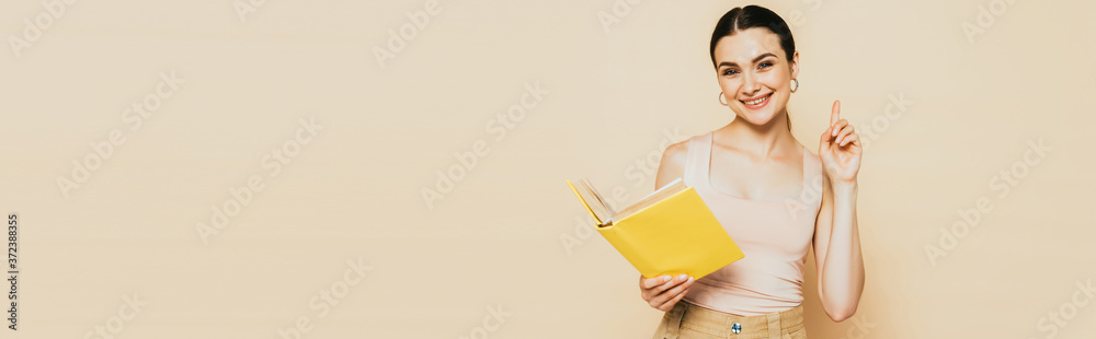 brunette young woman with yellow book showing idea gesture isolated on beige, panoramic shot brunette young woman with yellow book showing idea gesture isolated on beige, panoramic shot
