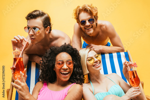 young shirtless men in sunglasses having fun with multicultural women sitting in deck chairs with cocktails isolated on yellow