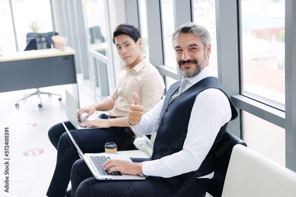 Smiling Senior Caucasian businessman Thumbs up and working with laptop in workspace at office. He sitting distance from asian staff and drinking a cup of hot coffee. New normal lifestyle concept. Smiling Senior Caucasian businessman Thumbs up and working with laptop in workspace at office. He sitting distance from asian staff and drinking a cup of hot coffee. New normal lifestyle concept.