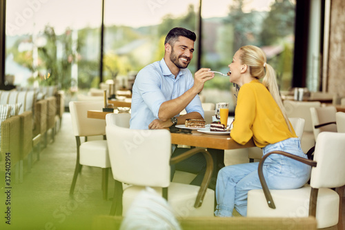 Cheerful man feeding his girlfriend with a cake in a cafe.