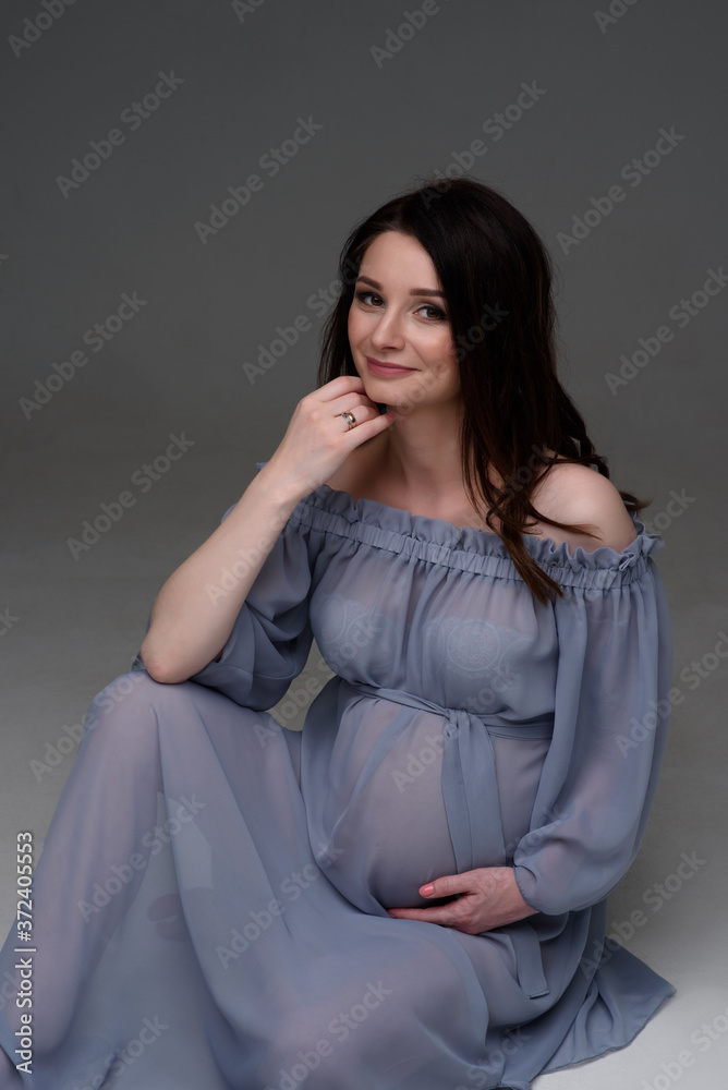 A young pregnant woman in a dress is sitting on the floor in the studio.