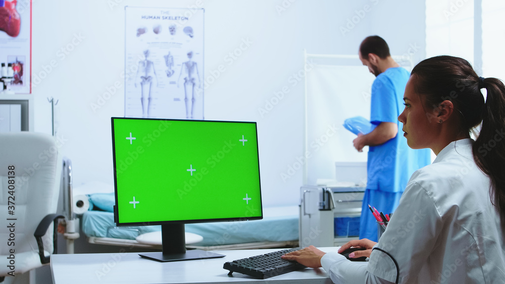 Doctor using computer with green screen display in hospital cabinet and assistant in the background. Medic in white coat working on monitor with chroma key in clinic cabinet to check patient diagnose.