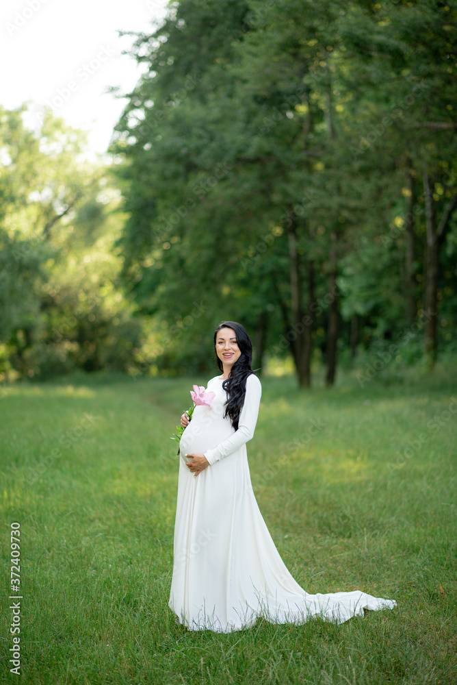Pregnant woman posing in a white dress on a background of green trees. Pregnant woman posing in a white dress on a background of green trees.