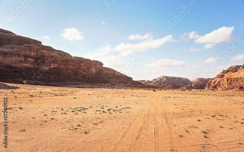Fototapeta Naklejka Na Ścianę i Meble -  Rocky massifs on red sand desert, bright cloudy sky in background, small 4wd vehicle at distance - typical scenery in Wadi Rum, Jordan